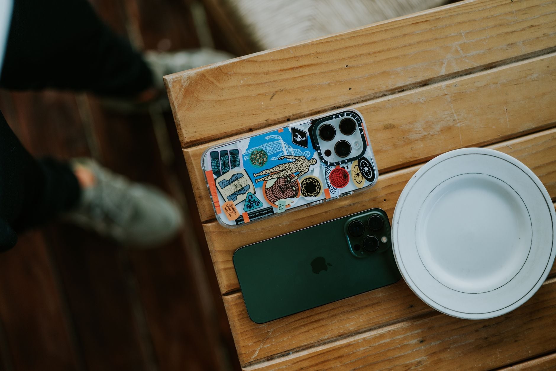 Tre smartphones med forskellige covers på træbord ved hvid tallerken, en med fisker-tema og en grøn variant.